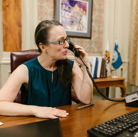 A female executive sits at a wooden desk, speaking on a landline phone in an office setting.