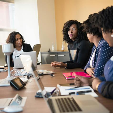 A group of people sitting at a conference table, engaged in discussion, with laptops and documents in front of them.