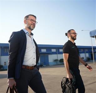 A man in a suit and a man in a black shirt and pants walking outdoors near an industrial blue building, both carrying bags and smiling.