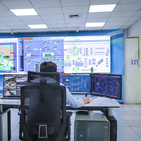 A person sits at a workstation with multiple monitors displaying complex system diagrams and controls in an intelligence operations center control room.