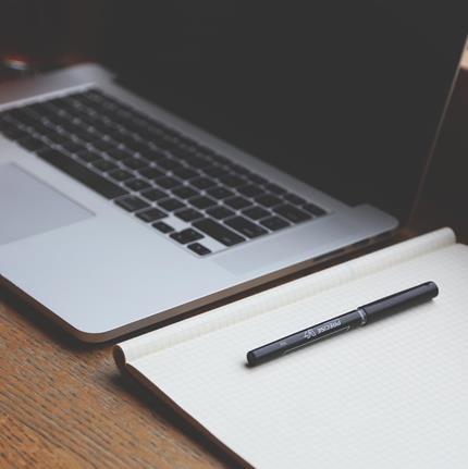 A wooden desk with a laptop, notebook and pen