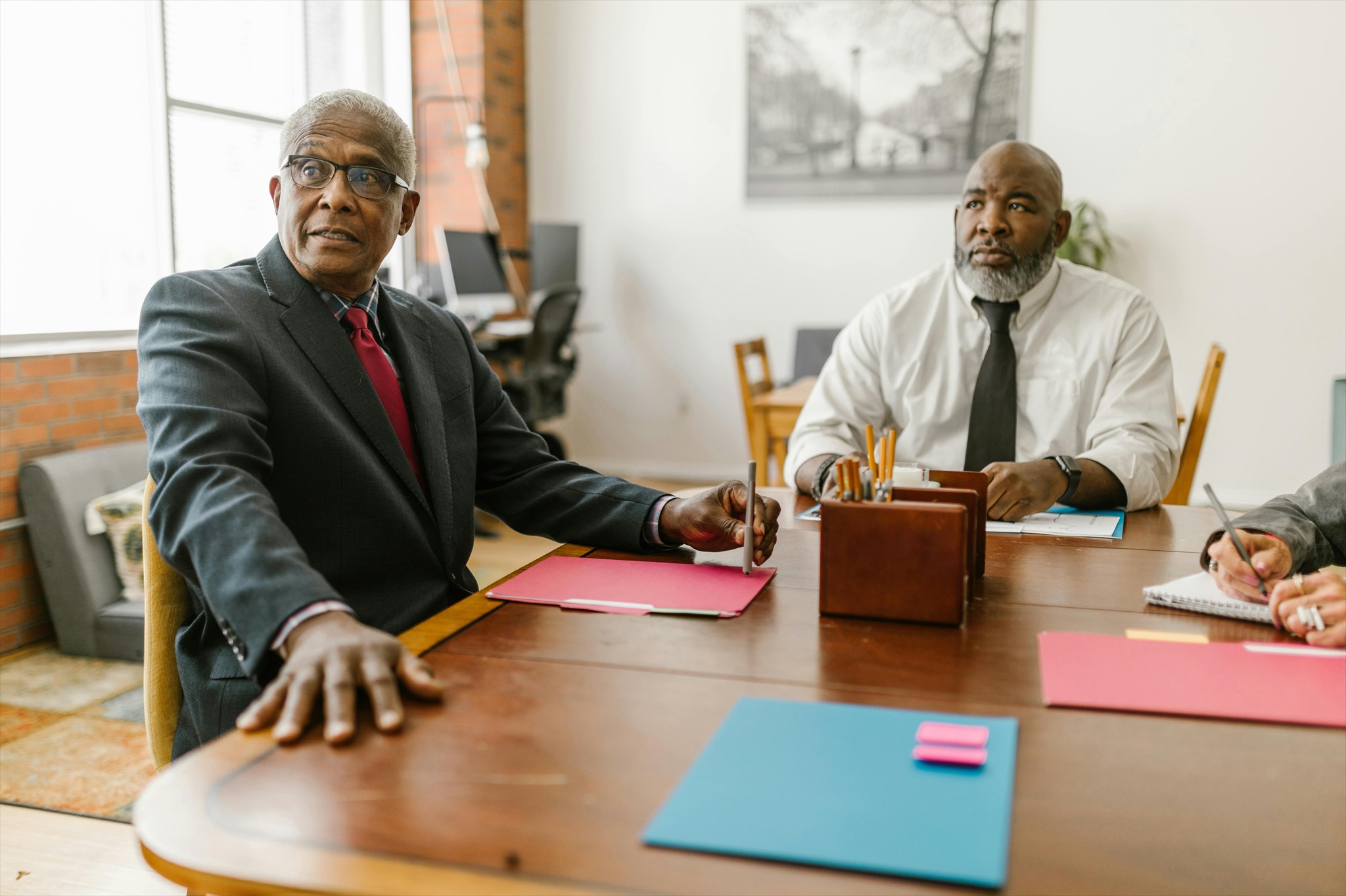 Two male executives in formal attire sit at a conference table with documents and supplies, appearing focused during a meeting.