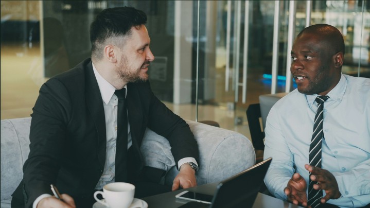 Two male executives in business attire engaged in a discussion while seated at a table with a laptop, notebook, and coffee cup.